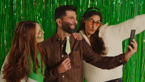 Studio-Shot-Of-Friends-Dressing-Up-With-Irish-Novelties-And-Props-Posing-For-Selfie-Celebrating-St-Patrick's-Day-Against-Green-Tinsel-Background